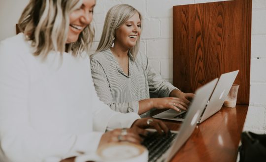 2 Women are sitting at a table typing on their laptop computers