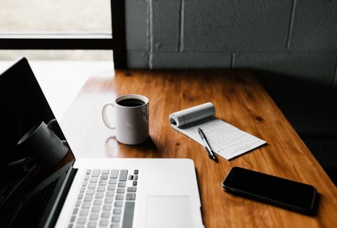 A laptop computer, phone, cup of coffee and a notepad are on a wooden desk