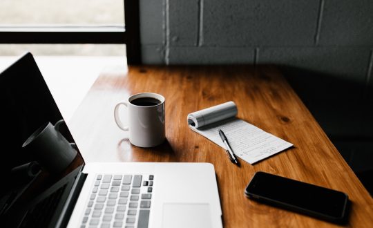 A laptop computer, phone, cup of coffee and a notepad are on a wooden desk