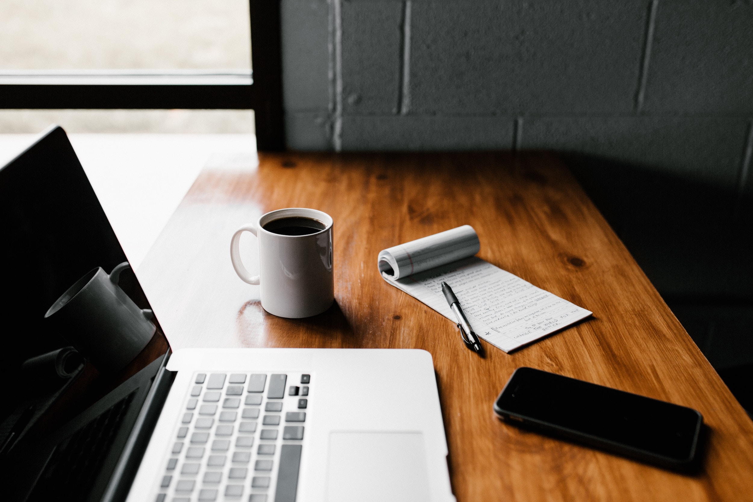 A laptop computer, phone, cup of coffee and a notepad are on a wooden desk