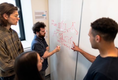 Men and woman are standing in front of a whiteboard discussing something